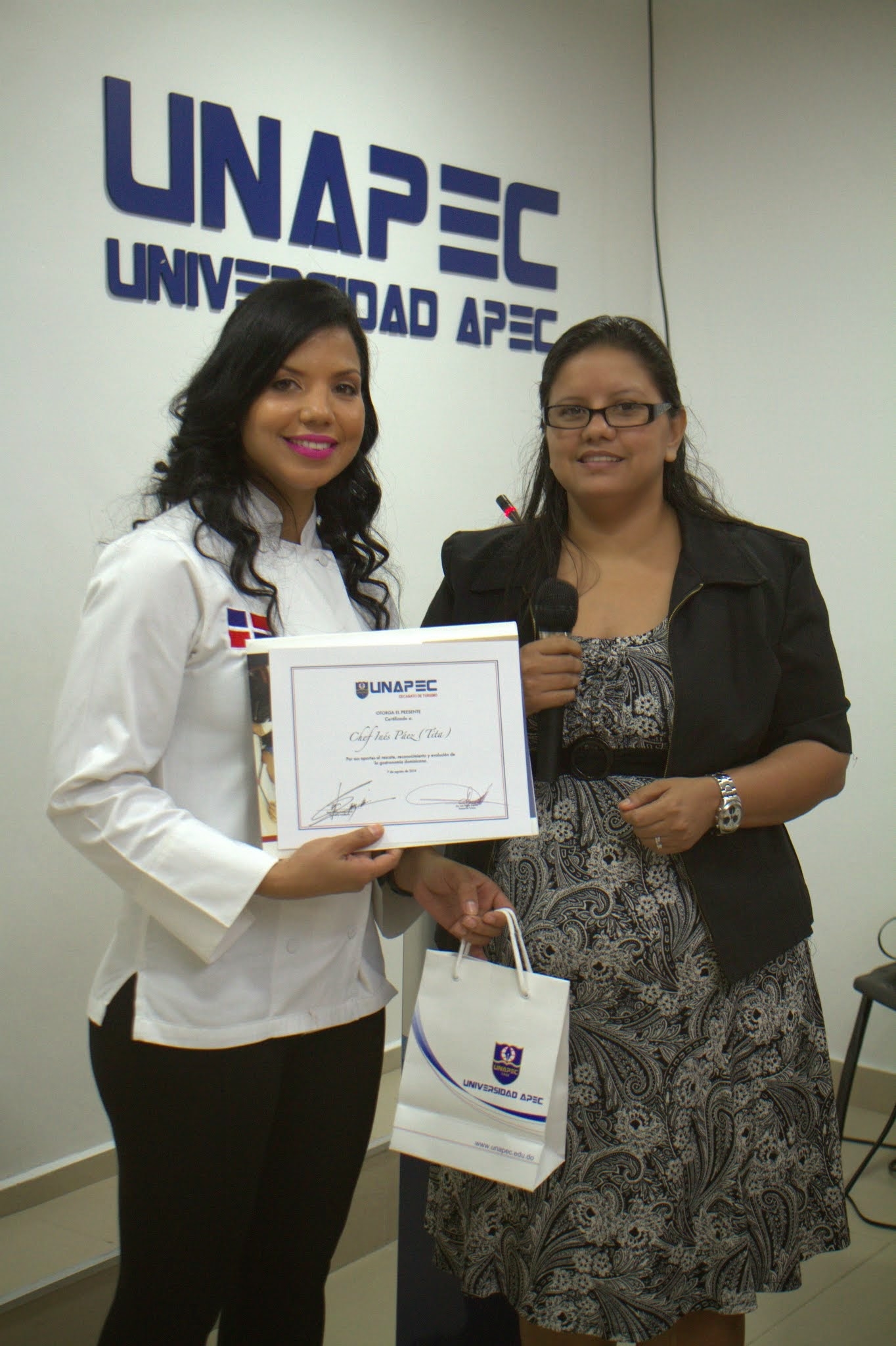 Inés Páez, recibiendo reconocimiento por parte de Yuderka Monción, Encargada del Laboratorio de Cocina de UNAPEC, en nombre de la Alta Casa de Estudios.